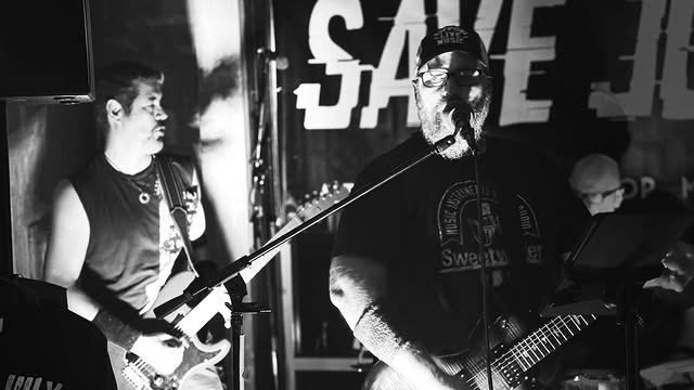 Black and white shot of the singer and guitarist in front of the Save July banner